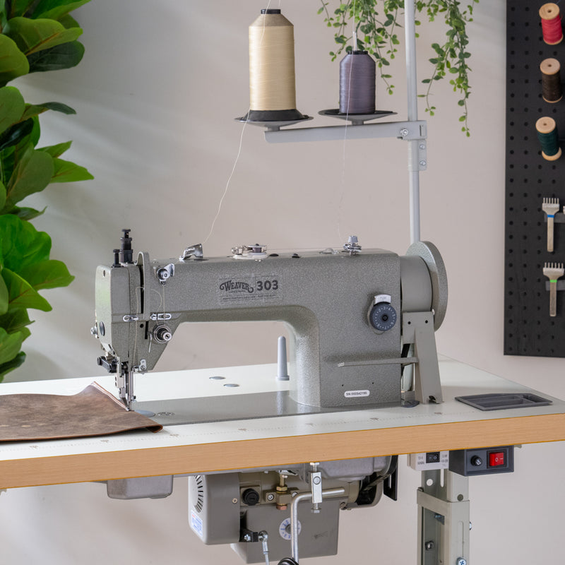 Weaver 303 leather sewing machine on work table with piece of brown leather and green plant in background.