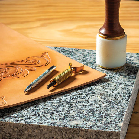 Leather tooling setup with swivel knife, beveler, mallet, and tooled floral design on veg-tan leather over marble slab.