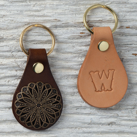 Two handmade leather key fobs, one dark brown with a decorative concho and one natural with a stamped "W" logo.